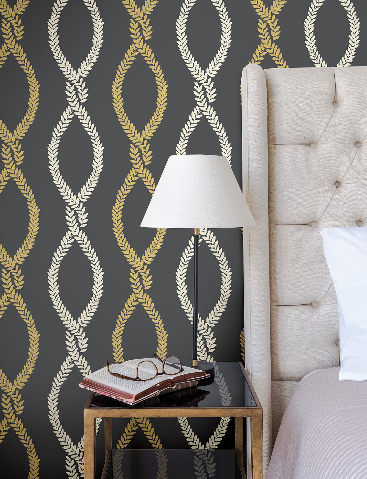 A beige tufted bed, black side table with books and glasses, and a white lamp sit against a dark wall covered in Laureate Vine Trellis Wallpaper by York Wallcoverings, featuring a gold and cream laurel wreath pattern.