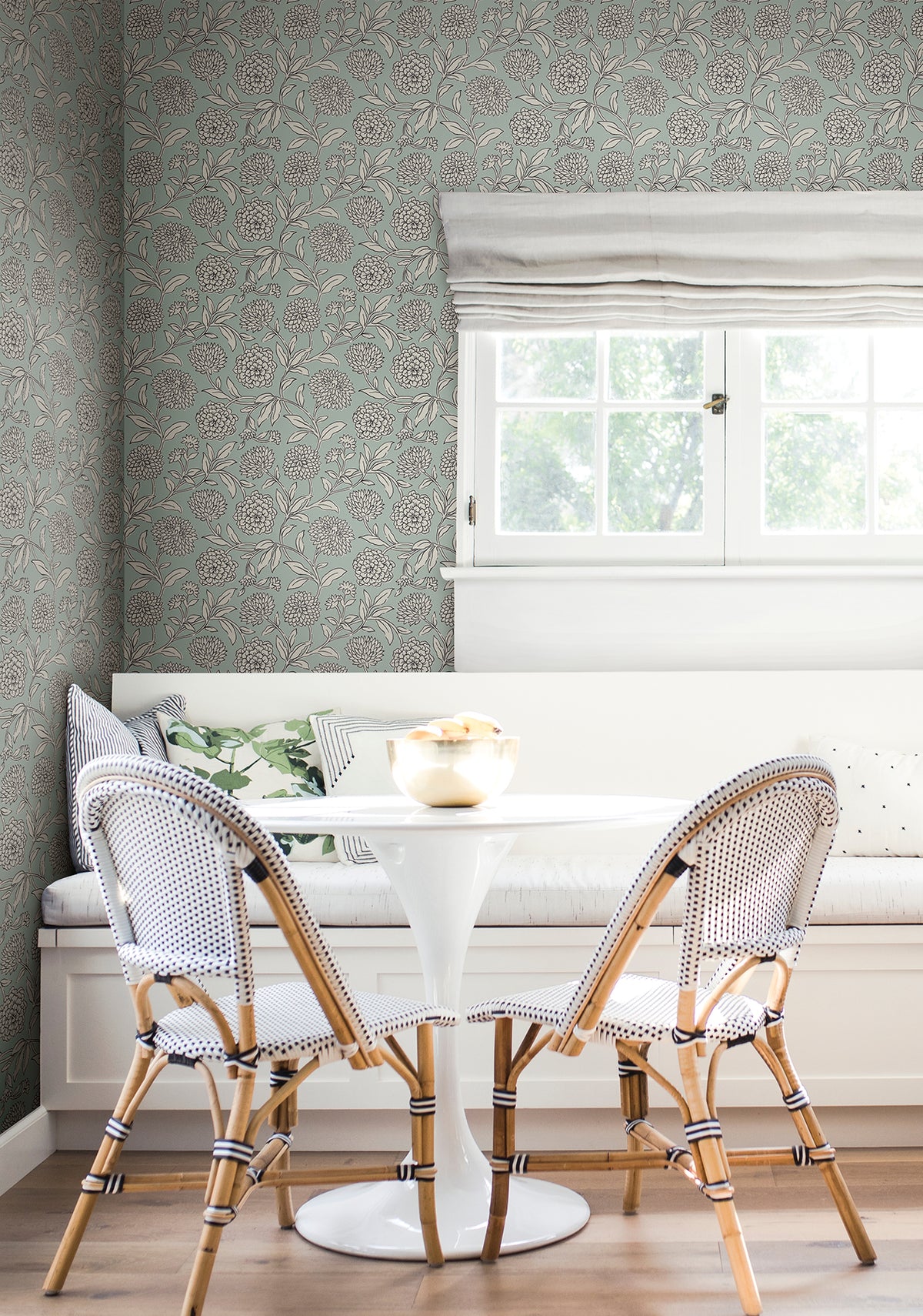A cozy breakfast nook with a round white table, two woven chairs, a built-in bench with cushions, and a window with a Roman shade letting in natural light is finished with York Wallcoverings’ Chatsworth Garden Floral Trail Wallpaper for added charm.