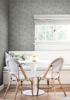 A cozy breakfast nook with a round white table, two woven chairs, a built-in bench with cushions, and a window with a Roman shade letting in natural light is finished with York Wallcoverings’ Chatsworth Garden Floral Trail Wallpaper for added charm.
