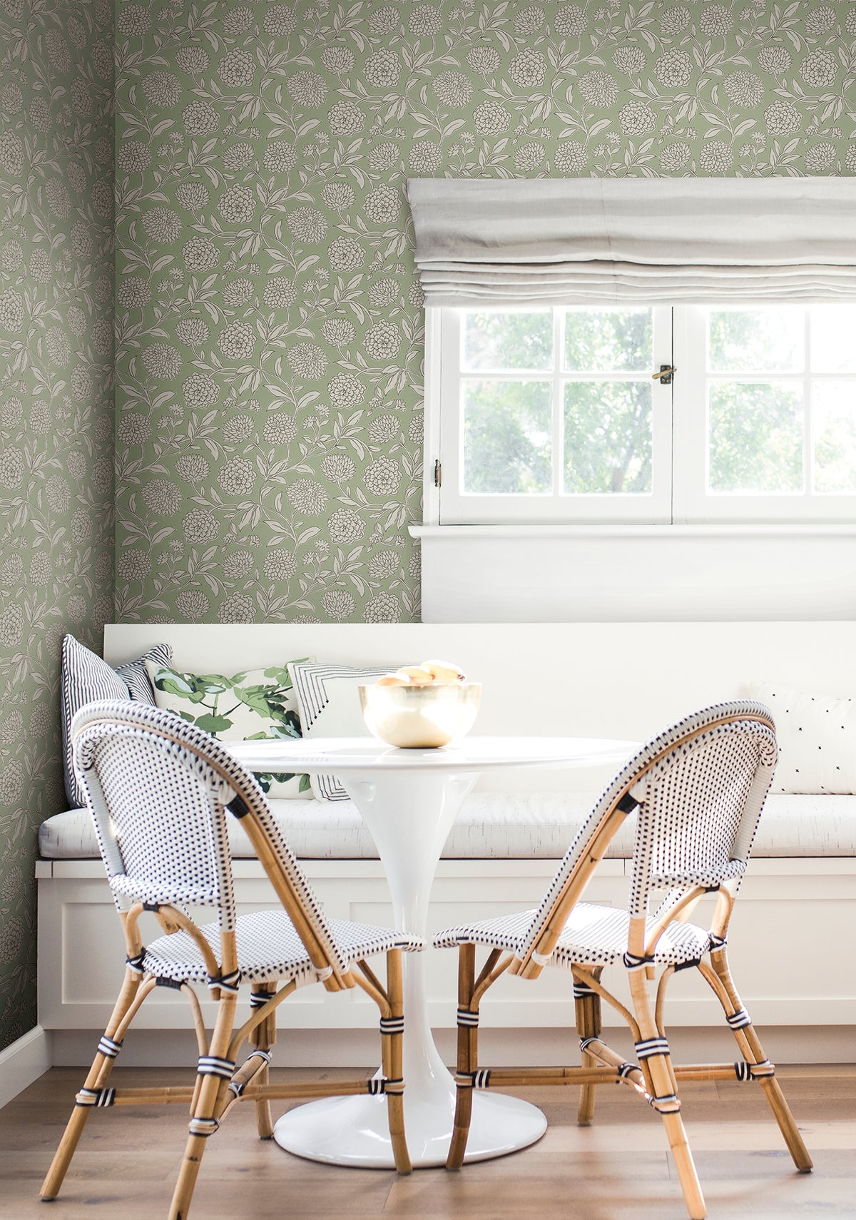 A cozy breakfast nook featuring a round white table, two woven chairs, a built-in bench with pillows, floral Chatsworth Garden Floral Trail Wallpaper by York Wallcoverings, and a window dressed with white roman shades letting in natural light.