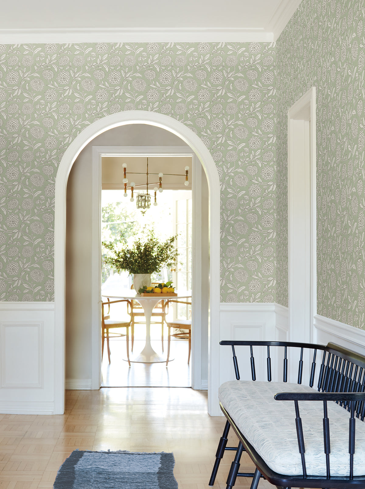 A hallway adorned with Chatsworth Garden Floral Trail Wallpaper by York Wallcoverings, featuring an arched doorway, black bench with white cushion, and a view into a dining area with a round table, wooden chairs, and a vase of flowers.
