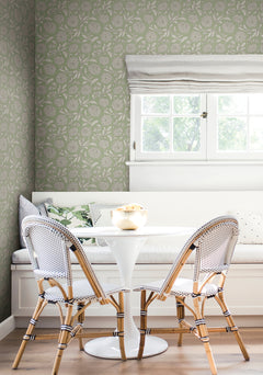 A small dining nook features a white round table, two woven chairs, and a built-in bench with pillows beneath a window. The walls are adorned with York Wallcoverings’ Chatsworth Garden Floral Trail Wallpaper in green. A bowl of fruit sits on the table.