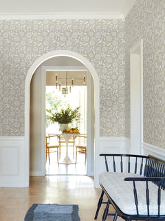 A bright hallway features Chatsworth Garden Floral Trail Wallpaper by York Wallcoverings, a black bench with a cushion, and an arched doorway that opens into a dining area with a round white table, wooden chairs, and a vase of greenery.