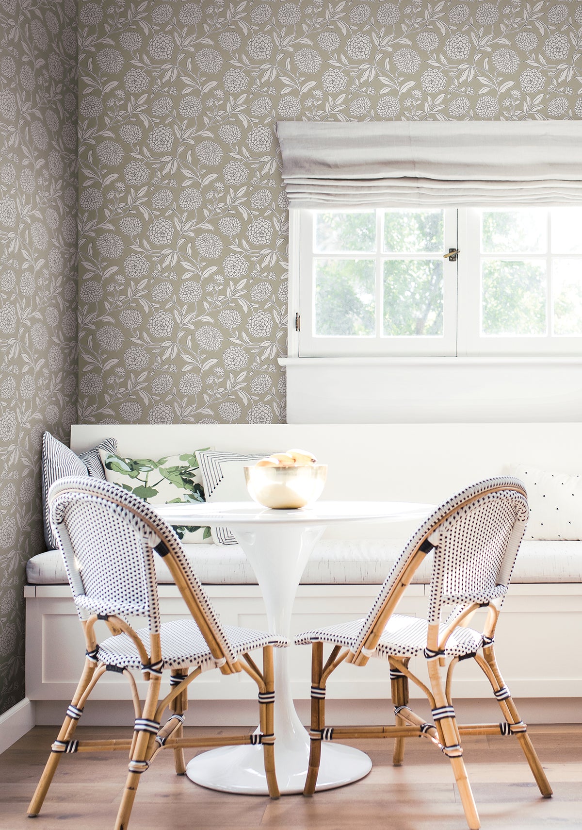 Bright, cozy breakfast nook with a round white table, two woven chairs, a built-in bench with cushions, and a window with white Roman shades letting in light. The space features Chatsworth Garden Floral Trail Wallpaper by York Wallcoverings.