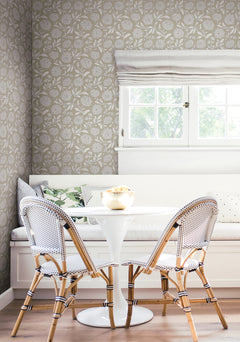 A bright breakfast nook features a white round table, two woven chairs, a built-in bench with cushions, and floral walls adorned with Chatsworth Garden Floral Trail Wallpaper by York Wallcoverings. Natural light streams in through a window with white blinds.