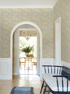 A hallway adorned with York Wallcoverings’ Chatsworth Garden Floral Trail Wallpaper in yellow and gray, featuring a black bench with a cushion, and an arched doorway that opens to a sunlit dining area with a round table, chairs, and a vase of greenery.