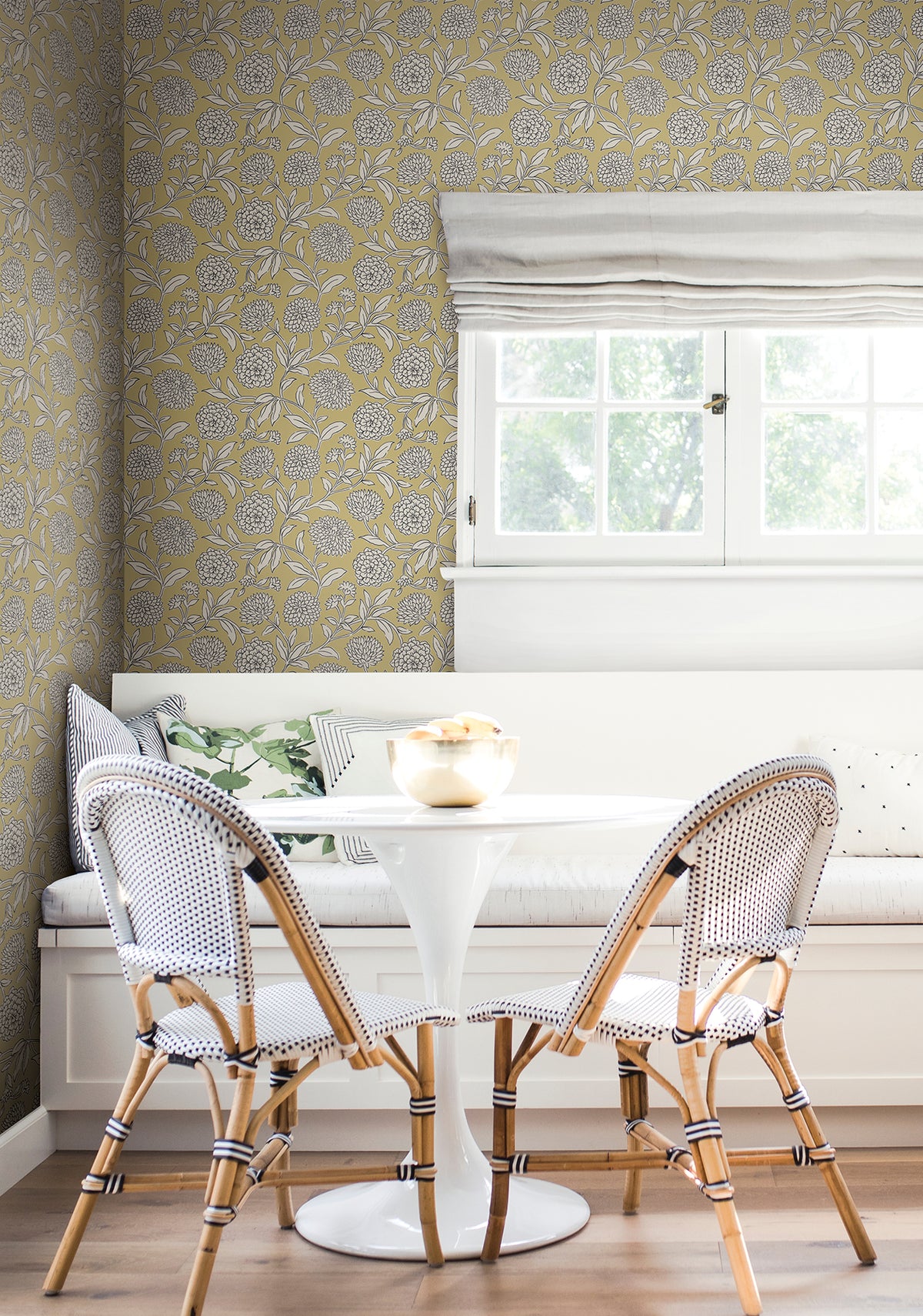 A bright nook featuring a white round table, two woven chairs, a built-in bench with pillows, and a window with a white shade that lets in natural light—all set against York Wallcoverings’ Chatsworth Garden Floral Trail Wallpaper.