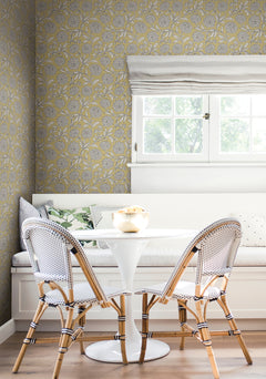 A cozy breakfast nook with a round white table, two wicker chairs, a built-in bench with cushions, and a window above. The walls are covered in Chatsworth Garden Floral Trail Wallpaper by York Wallcoverings.