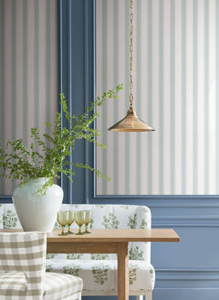 A dining area featuring a wooden table, patterned upholstered chairs, a white vase with greenery, green glasses, and a brass pendant light set against York Wallcoverings’ Tilford Awning Stripe Wallpaper with blue trim.