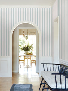 A hallway adorned with Tilford Awning Stripe Wallpaper by York Wallcoverings features an arched doorway opening to a dining area with a round table, chairs, a vase of flowers, and a chandelier. In the foreground, there is a black bench with a cushion and a rug.