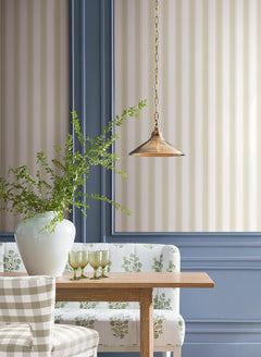 A dining nook featuring a wooden table, patterned chairs, a floral bench, green glasses, a white vase with greenery, and a brass pendant light is set against blue-trimmed walls covered in York Wallcoverings’ Tilford Awning Stripe Wallpaper.