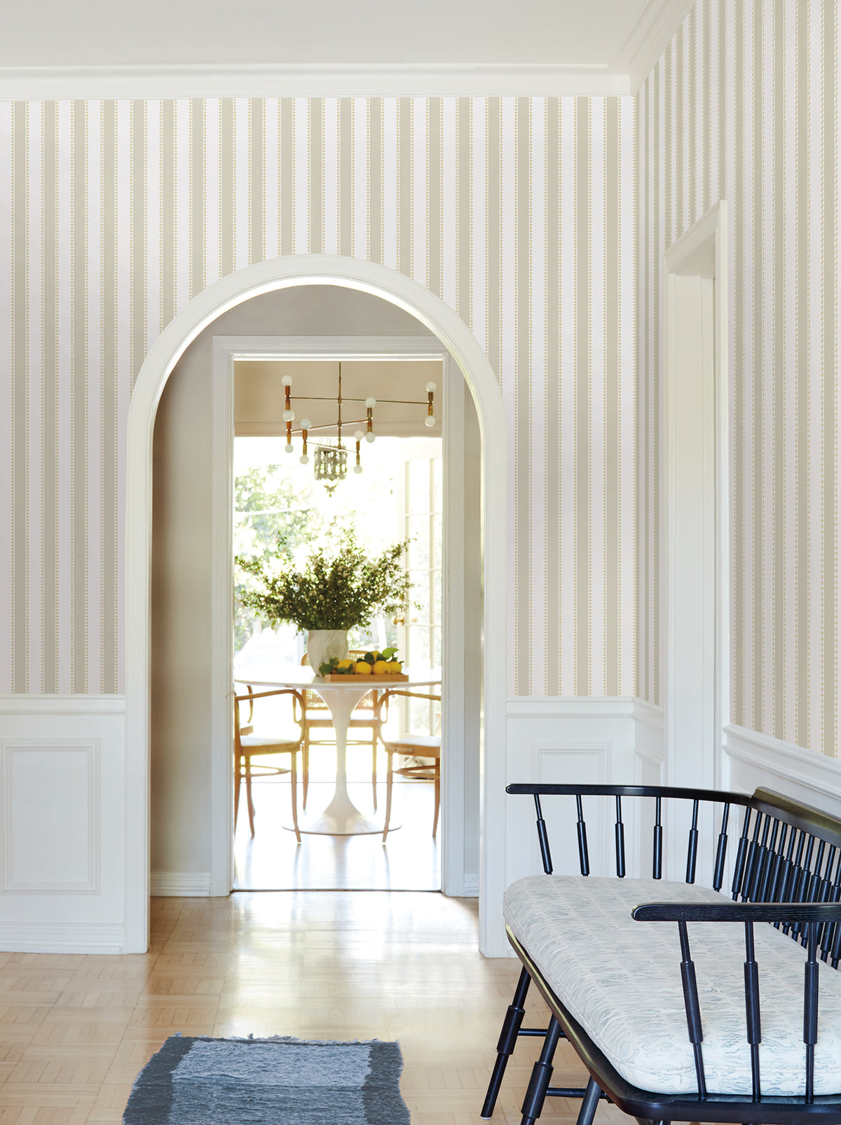 A hallway featuring Tilford Awning Stripe Wallpaper by York Wallcoverings, a black bench topped with a cushion, and a blue rug leads through an arched doorway to a bright dining area with a round table, chairs, and a large vase of flowers.