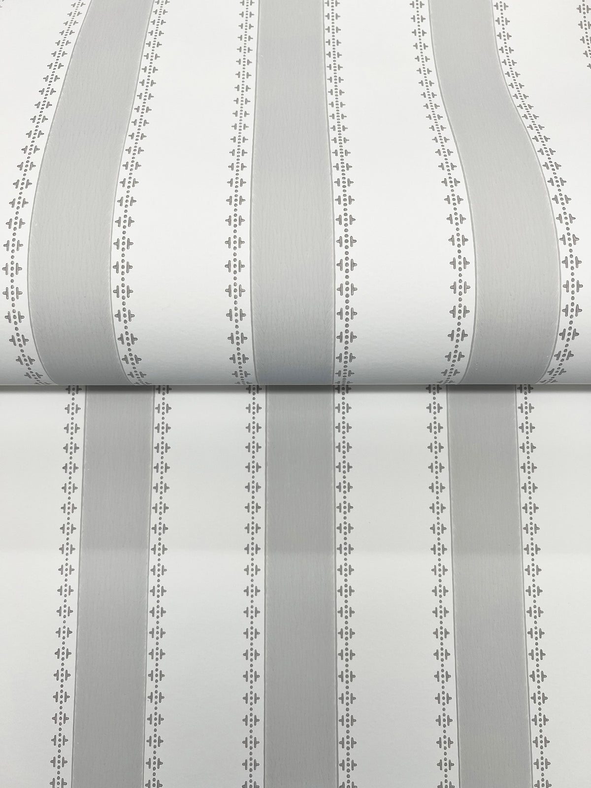 A close-up of the Tilford Awning Stripe Wallpaper by York Wallcoverings, featuring vertical gray and white stripes bordered by small, repeating diamond and dot patterns. The wallpaper is shown partially unrolled on a flat surface.