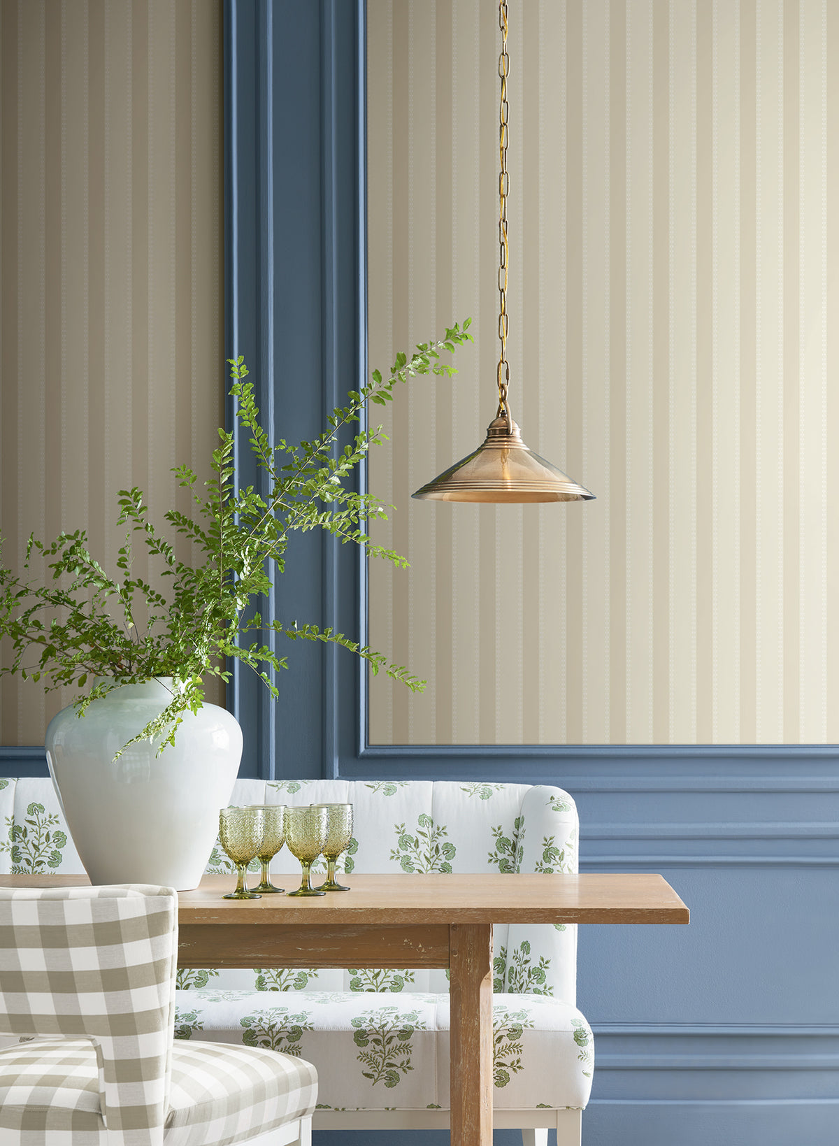 A gold pendant light hangs over a wooden table with a white vase of green branches, three green glasses, and patterned bench seating against walls adorned with York Wallcoverings’ Tilford Awning Stripe Wallpaper.