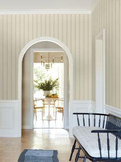 A hallway featuring York Wallcoverings’ Tilford Awning Stripe Wallpaper in beige, accented by white trim and an arched doorway opening into a sunlit dining room. In the foreground, a black bench with a cushion sits atop a small rug.