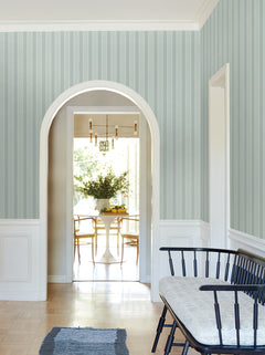 A hallway adorned with York Wallcoverings’ Tilford Awning Stripe Wallpaper in blue features a black bench with a cushion and a rug, leading through an arched doorway to a bright dining room with a round white table, chairs, and a vase of greenery.