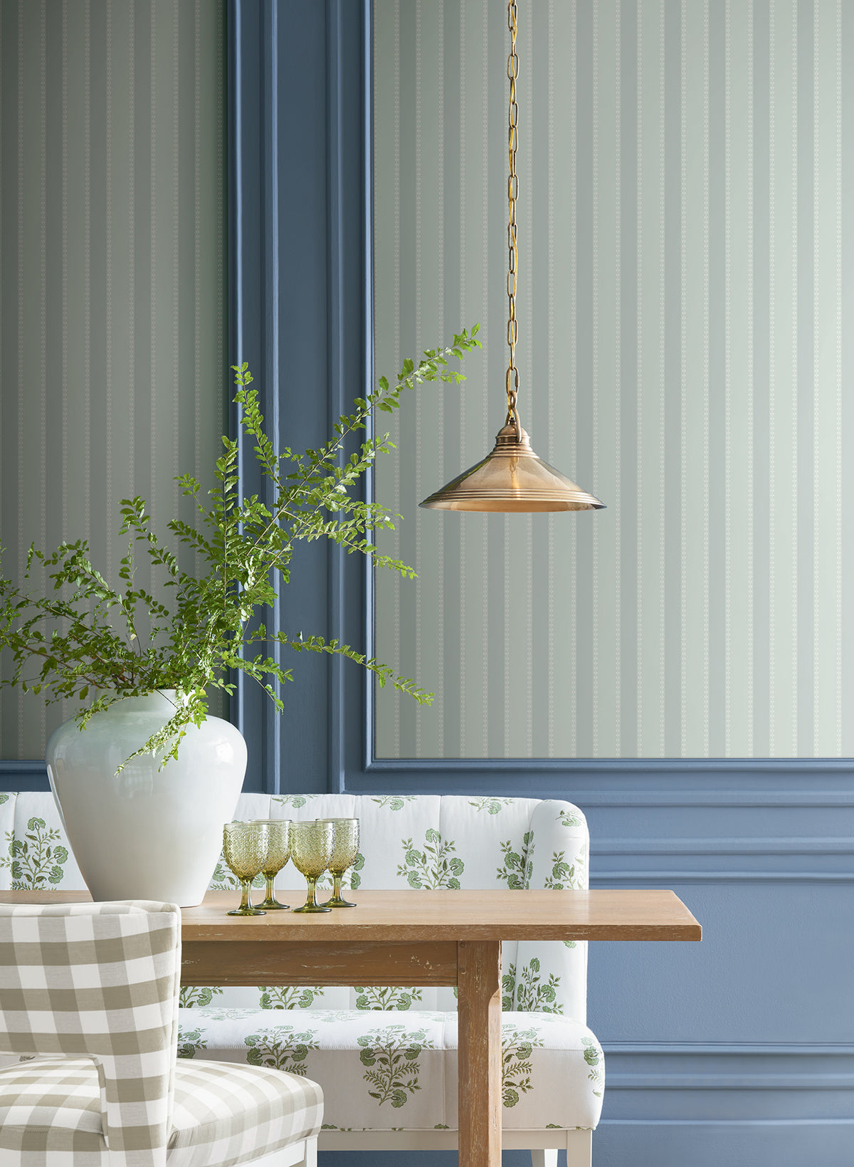 A wooden table with a white floral bench and checkered chair, a large white vase filled with green branches, three green glasses, and a brass pendant light, set against blue panel walls accented by Tilford Awning Stripe Wallpaper from York Wallcoverings.