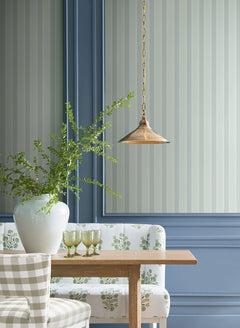 A wooden table with a white floral bench and checkered chair, a large white vase filled with green branches, three green glasses, and a brass pendant light, set against blue panel walls accented by Tilford Awning Stripe Wallpaper from York Wallcoverings.