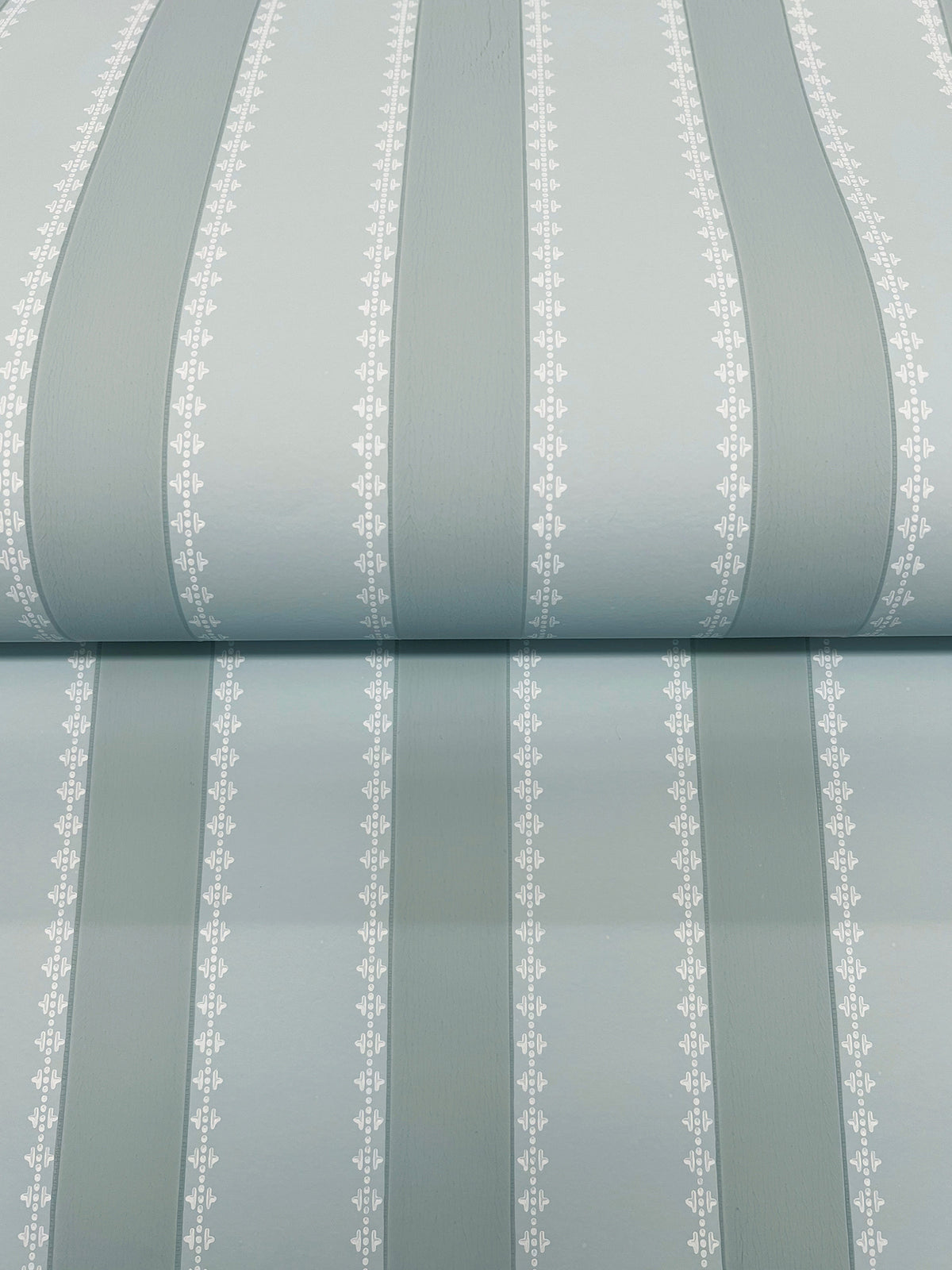 The image shows York Wallcoverings’ Tilford Awning Stripe Wallpaper, featuring light blue with wide, vertical sage-green stripes and thin white lines accented by small leaf patterns on each side. The wallpaper is rolled out with a visible horizontal seam across the center.