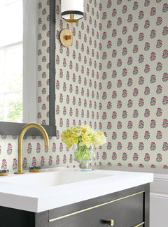 A bathroom featuring Sylvie Floral Wallpaper by York Wallcoverings, a white sink with a gold faucet, a black vanity, a round wall sconce with a gold base, a large mirror, and a glass vase of yellow roses. A window is visible on the left.