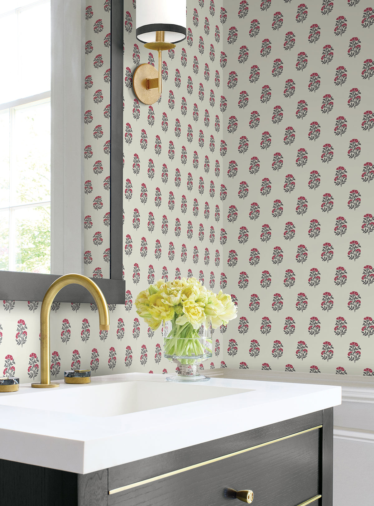 A bathroom features Sylvie Floral Wallpaper by York Wallcoverings, a white sink with a gold faucet, a black vanity, a vase of yellow flowers, a window, and a wall sconce with a gold base and white shade.