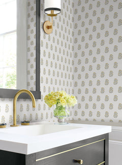 A bathroom featuring York Wallcoverings’ Sylvie Floral Wallpaper, a gold faucet, a glass vase with yellow flowers on the counter, a rectangular mirror, and a gold and black wall sconce beside a window.