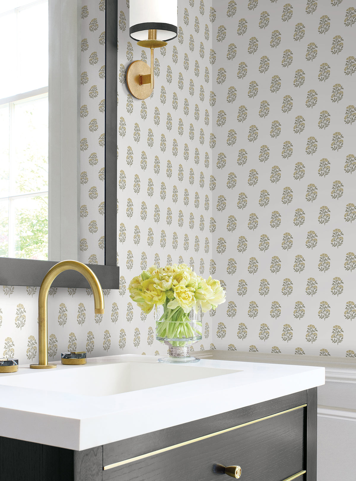 A modern bathroom features York Wallcoverings’ Sylvie Floral Wallpaper, gold accents, a black vanity with a white countertop, and a glass vase filled with yellow flowers next to a gold faucet. Sunlight pours in through a nearby window.
