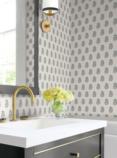 Elegant bathroom featuring Sylvie Floral Wallpaper by York Wallcoverings, a gold faucet, black vanity with gold accents, white countertop, wall sconce, and a glass vase holding yellow flowers beside a large mirror and window.
