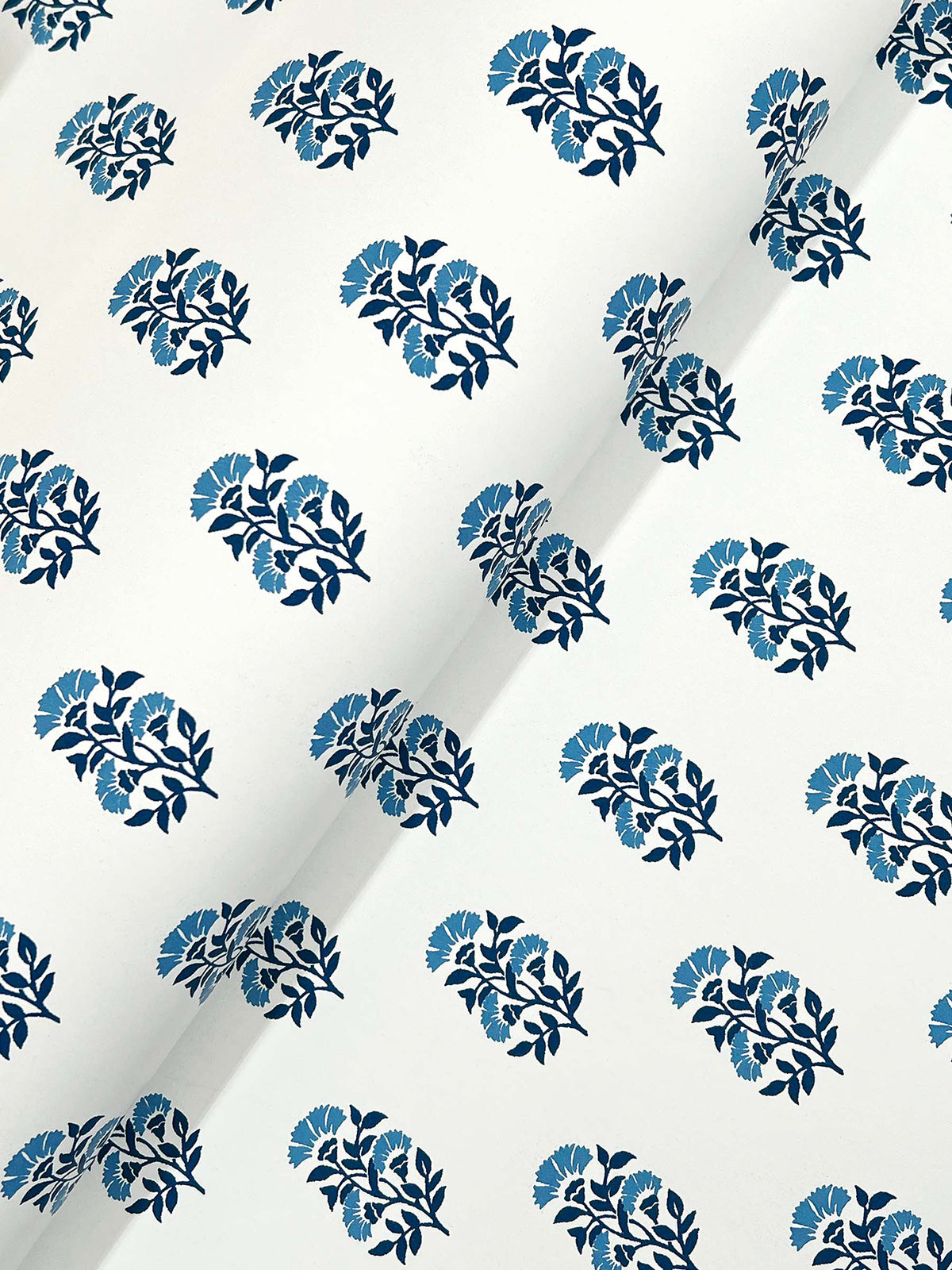 The Sylvie Floral Wallpaper by York Wallcoverings features a white surface adorned with a repeating pattern of dark blue leaves and light blue flowers, uniformly arranged across the material. A diagonal crease is visible in the image.