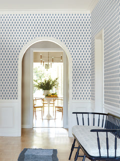 A hallway adorned with Sylvie Floral Wallpaper by York Wallcoverings features an arched doorway that leads to a dining area with a round table, chairs, large windows, and a vase of flowers; a black bench is positioned against the wall.