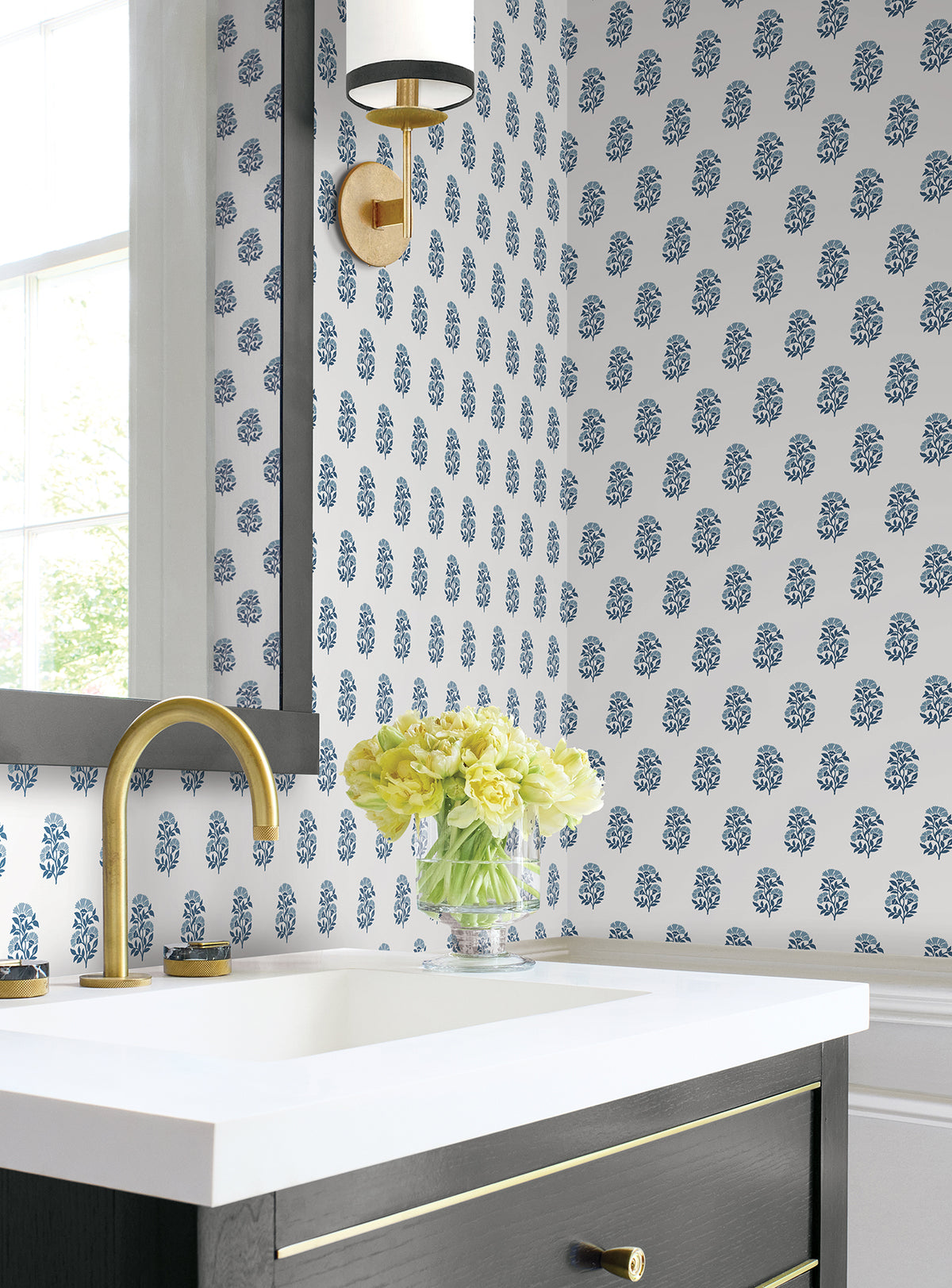 A modern bathroom featuring a white sink, gold faucet, and black vanity is brightened by a vase of yellow flowers. The wall is adorned with York Wallcoverings’ Sylvie Floral Wallpaper in blue, while a round light fixture with gold accents is mounted above the sink.