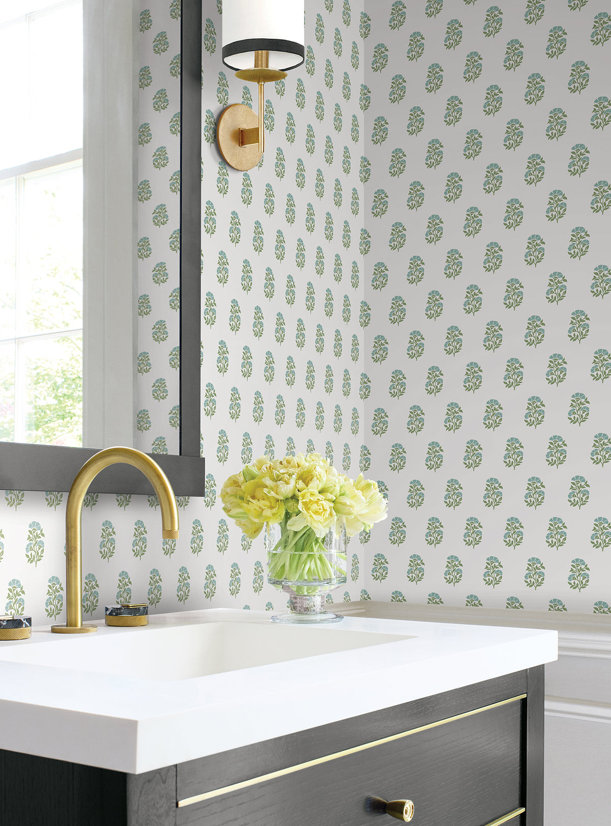 Bathroom featuring York Wallcoverings’ Sylvie Floral Wallpaper, a gold faucet, a black vanity with gold accents, and a vase of yellow flowers on the counter beneath a wall sconce and window.