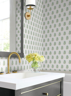 Bathroom featuring York Wallcoverings’ Sylvie Floral Wallpaper, a gold faucet, a black vanity with gold accents, and a vase of yellow flowers on the counter beneath a wall sconce and window.