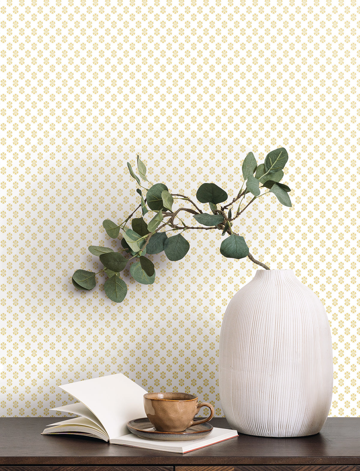 A white ribbed vase with green branches, an open notebook, and a brown cup and saucer sit on a dark wooden table against a wall covered in York Wallcoverings’ Brioni Floral Stamp Wallpaper.