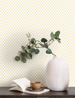 A white ribbed vase with green branches, an open book, and a brown cup and saucer sit on a dark wooden table in front of a wall adorned with Brioni Floral Stamp Wallpaper by York Wallcoverings.