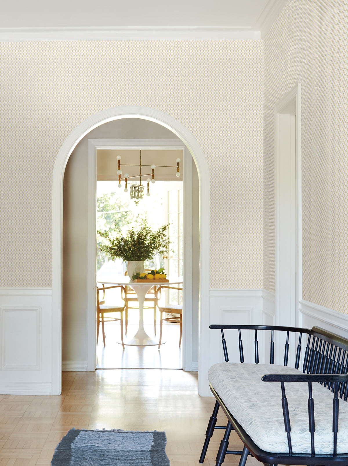 Brioni Floral Stamp Wallpaper from York Wallcoverings adds subtle pattern to the bright hallway with an arched doorway leading to the dining area. A black bench with a cushioned seat sits on the right atop a blue rug, while sunlight fills the room and a vase of greenery decorates the dining table.