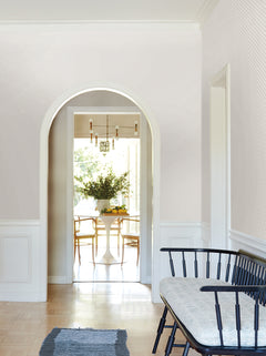 A sunlit dining area with a round table and wooden chairs is visible through an archway adorned with Brioni Floral Stamp Wallpaper by York Wallcoverings; in the foreground, there’s a black bench with a cushion and a blue rug on a light wood floor.