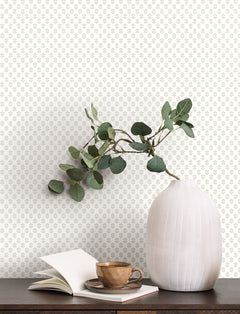 A ribbed white vase with green branches sits on a dark table next to an open book and a brown cup. The background is adorned with the Brioni Floral Stamp Wallpaper by York Wallcoverings, showcasing a delicate, repeating light green floral pattern.