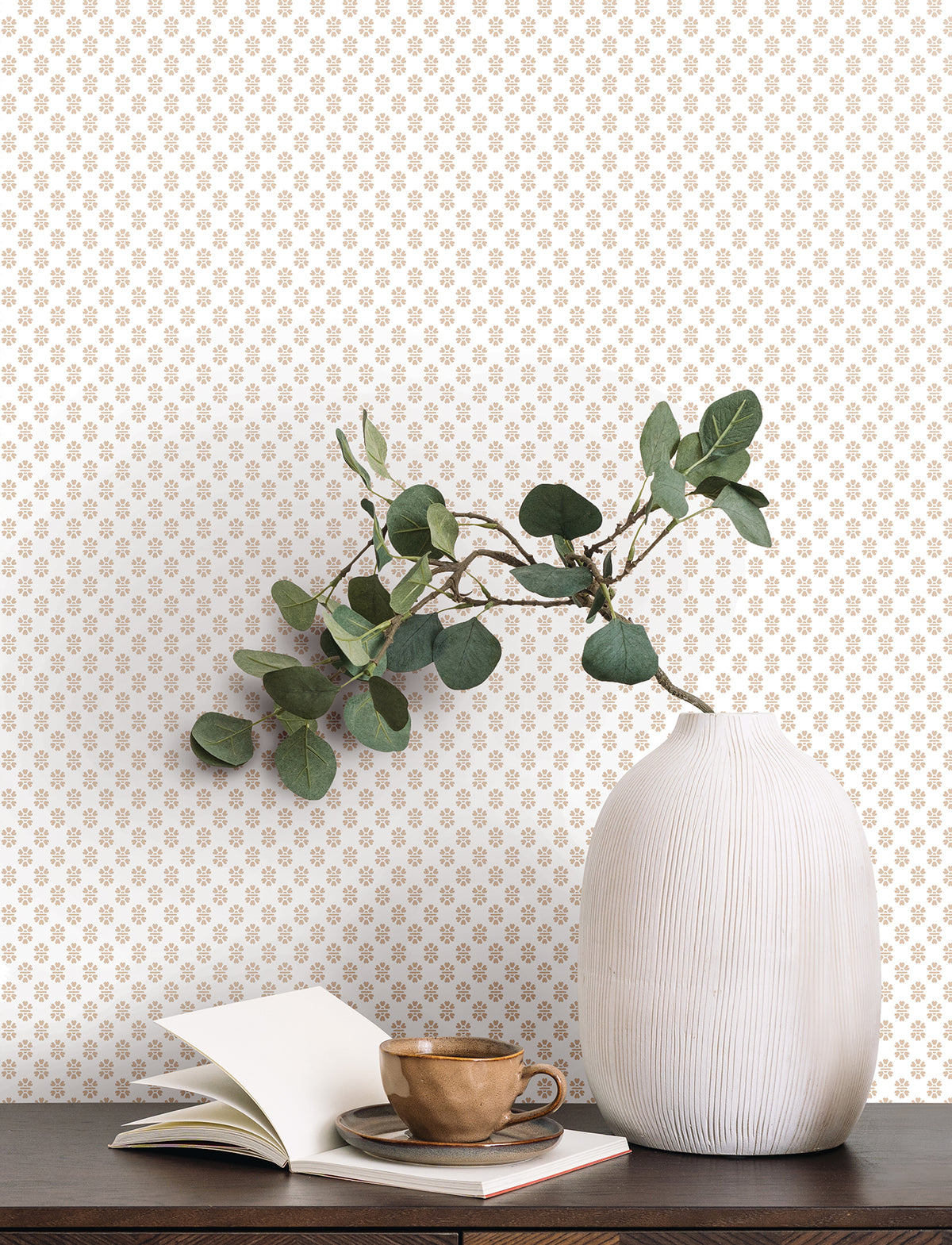 A white ribbed vase with green leaves sits on a dark wooden surface next to an open book and a brown cup and saucer. The background features York Wallcoverings’ Brioni Floral Stamp Wallpaper, showcasing a small, repeating gold floral pattern on a white wall.