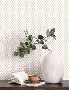 A white ribbed vase with eucalyptus branches sits on a dark table next to an open book and a brown cup and saucer. The background features York Wallcoverings’ Brioni Floral Stamp Wallpaper in white with a small, repeating tan floral pattern.