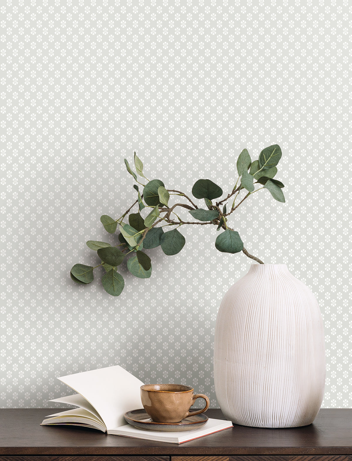 A white textured vase with green eucalyptus branches sits on a dark table next to an open notebook and a brown ceramic cup and saucer. In the background, the wall is adorned with Brioni Floral Stamp Wallpaper by York Wallcoverings in a light gray pattern.