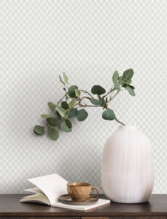 A white textured vase with green eucalyptus branches sits on a dark table next to an open notebook and a brown ceramic cup and saucer. In the background, the wall is adorned with Brioni Floral Stamp Wallpaper by York Wallcoverings in a light gray pattern.