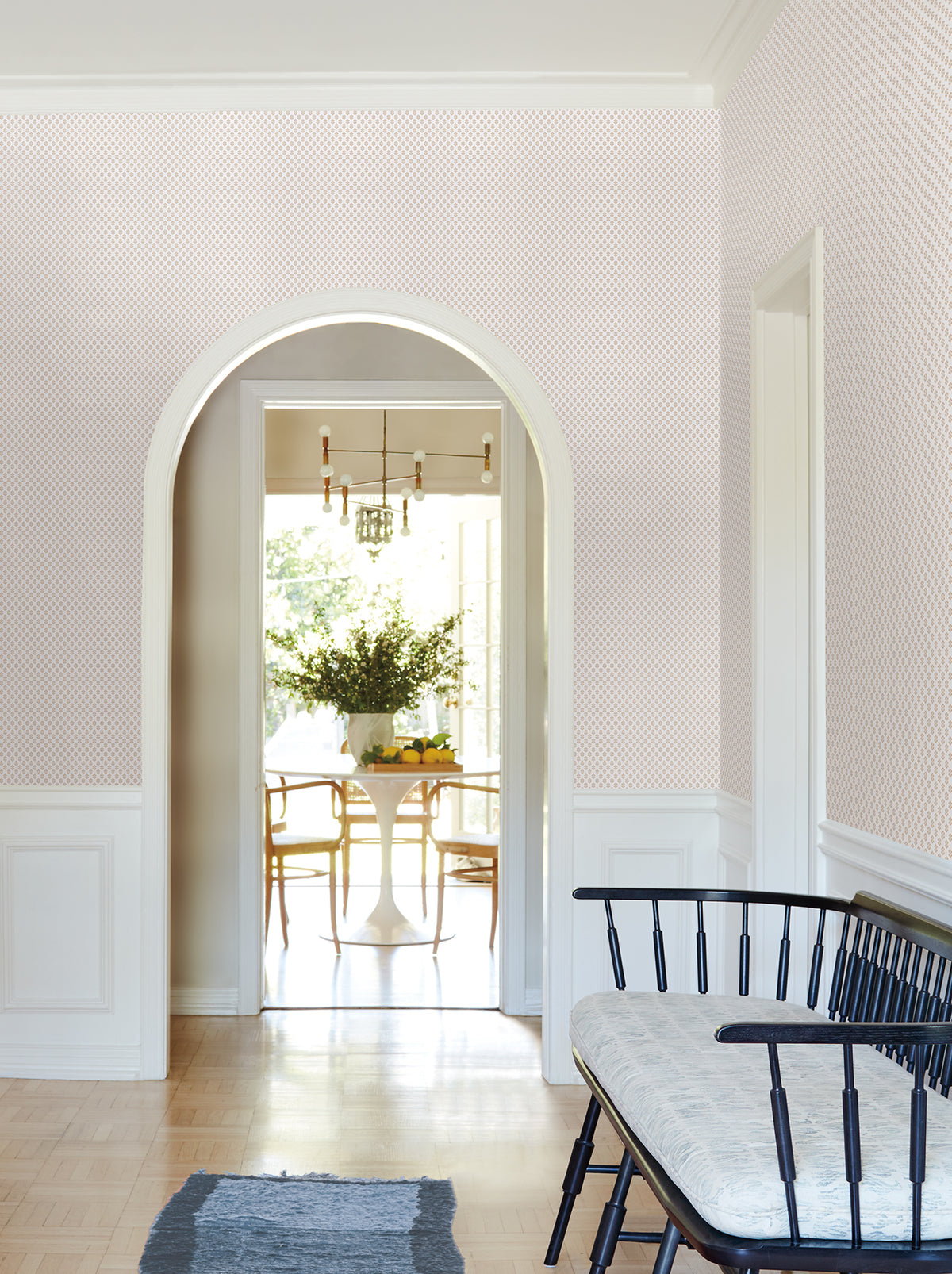 A bright hallway adorned with York Wallcoverings’ Brioni Floral Stamp Wallpaper and an arched doorway leads to a dining area featuring a round table, wooden chairs, a chandelier, and a vase of flowers. In the foreground, a black bench with a cushion adds an inviting touch.