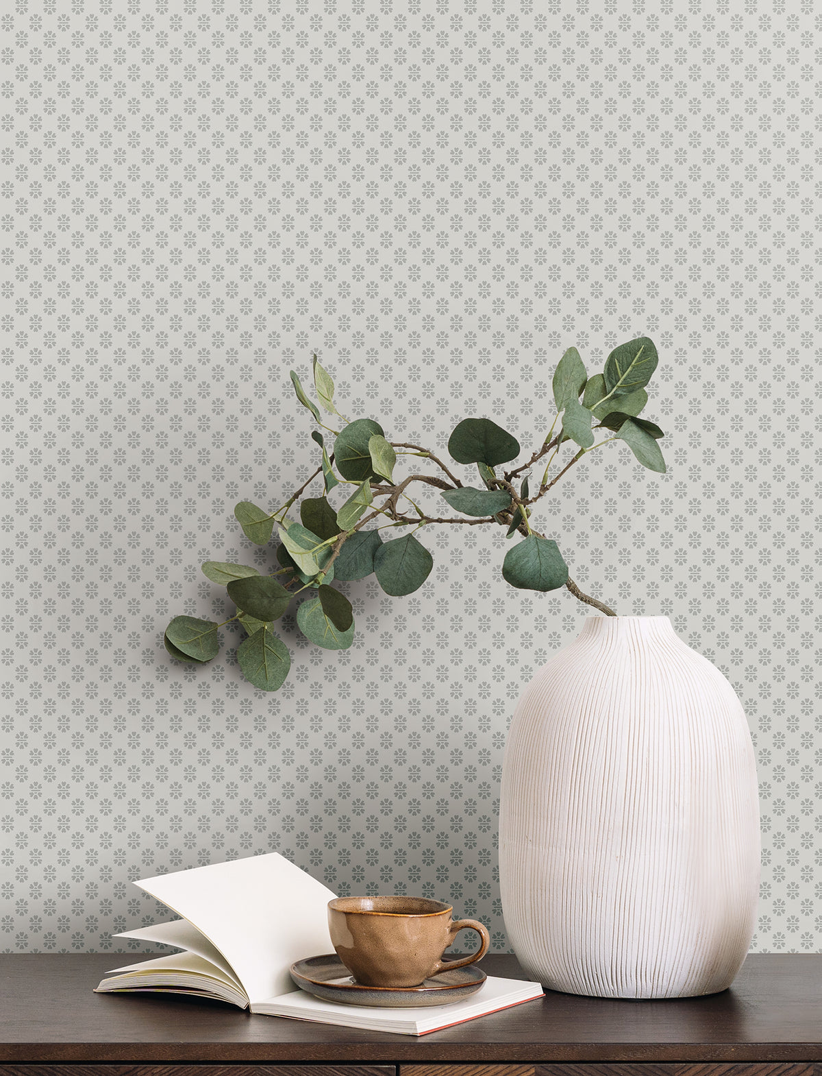 A white vase with green eucalyptus branches sits on a wooden table next to a brown cup and an open notebook. The background showcases York Wallcoverings’ Brioni Floral Stamp Wallpaper, featuring subtle, small grey floral patterns.