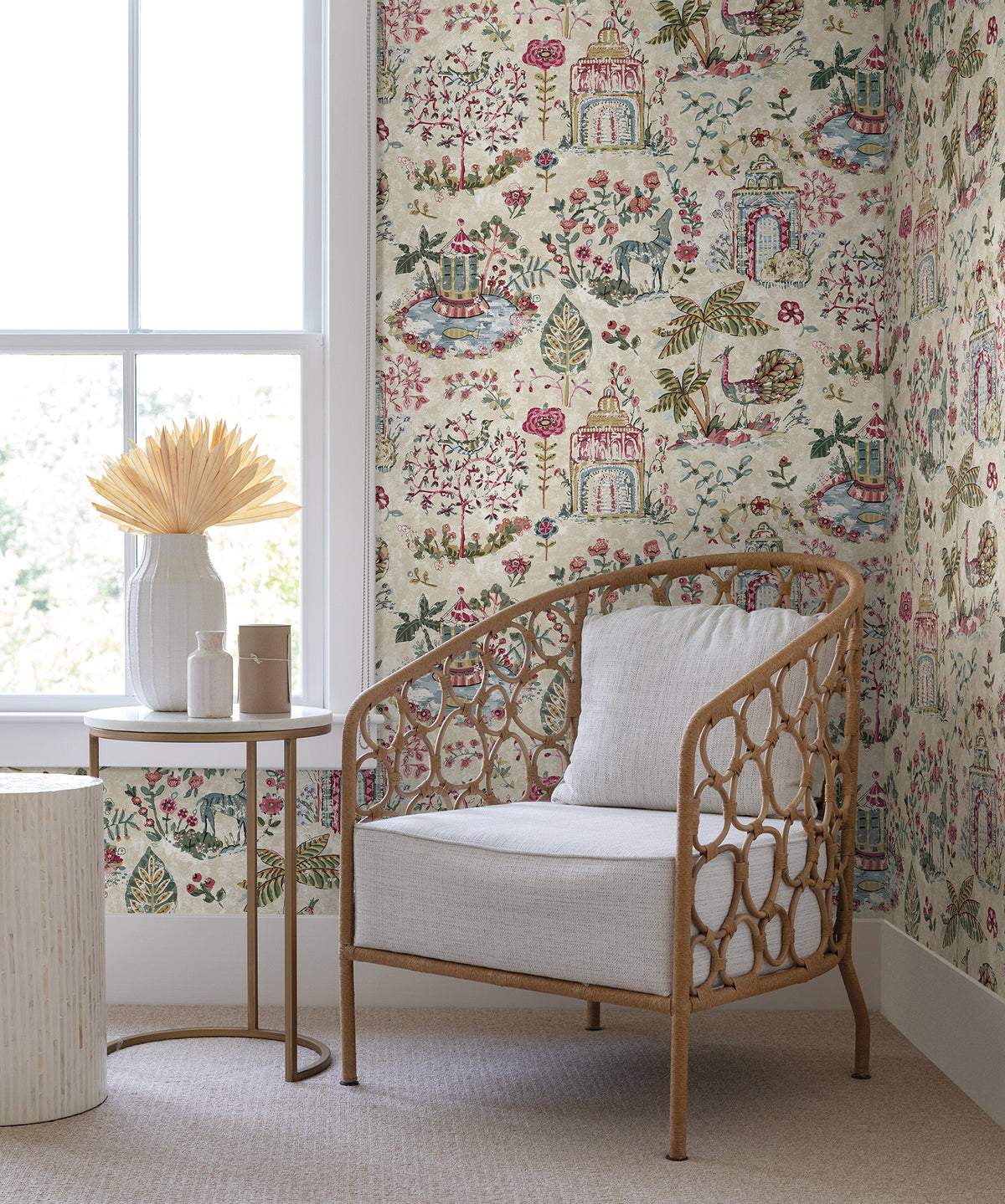 A cozy rattan chair with a white cushion sits beside a small round table adorned with vases and dried flowers, set against York Wallcoverings’ Parc Garden Dreams Wallpaper near a large window bathed in natural light.