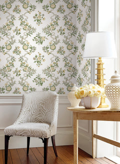 A chair with a light animal print sits beside a wooden table holding a lamp, floral arrangement, and decorative jar, set against a wall adorned with York Wallcoverings’ Genevieve Floral Trail Wallpaper. Sunlight streams in from the window.