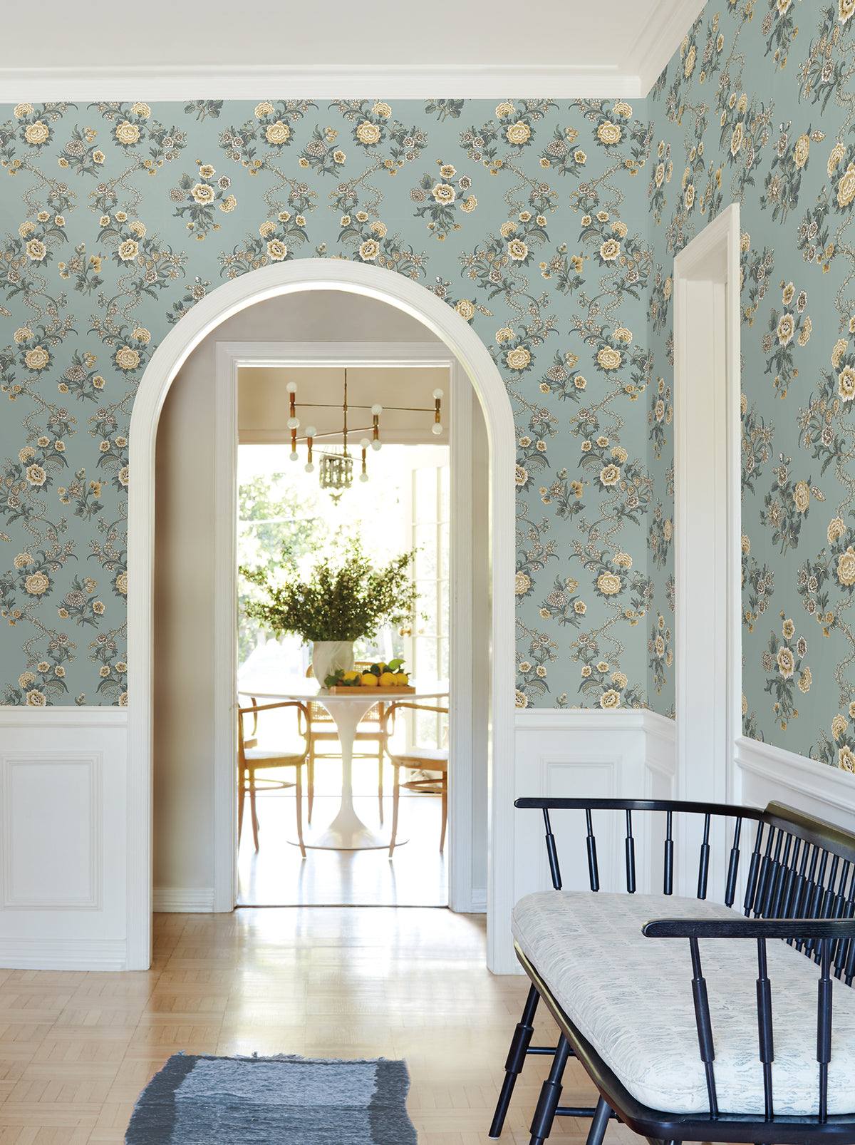 A hallway adorned with York Wallcoverings’ Genevieve Floral Trail Wallpaper in blue and white trim features an arched doorway leading to a bright dining area with a round table, chairs, a vase of flowers, and modern light fixtures. A bench sits against the wall in the foreground.