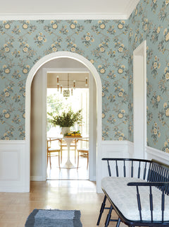 A hallway adorned with York Wallcoverings’ Genevieve Floral Trail Wallpaper in blue and white trim features an arched doorway leading to a bright dining area with a round table, chairs, a vase of flowers, and modern light fixtures. A bench sits against the wall in the foreground.