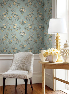 A cozy corner featuring a patterned chair and a wooden table topped with a gold lamp, white ceramic decor, and a bouquet of yellow roses. The background is adorned with York Wallcoverings’ Genevieve Floral Trail Wallpaper in blue and framed by crisp white trim, while sunlight pours in through the window on the right.