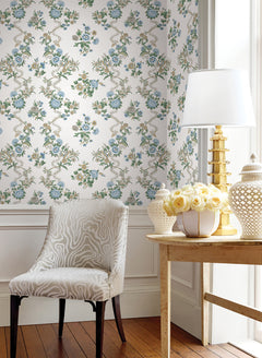 A stylish room corner featuring a patterned chair and a wooden table topped with yellow roses, white decorative accents, and a gold lamp. The wall is adorned with York Wallcoverings’ Genevieve Floral Trail Wallpaper in white with blue floral motifs, while sunlight pours in from the right.